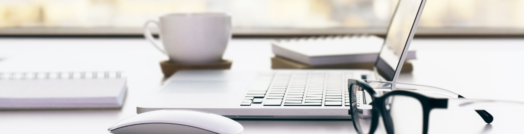 Desk surface with a laptop, a cup of coffee, a mouse, an open notebook and a pair of reading glasses